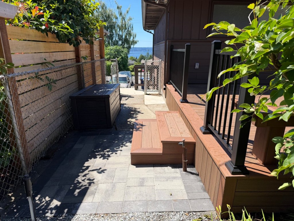 The side of the finished deck, with a composite step down onto the paver patio and black metal railing above.