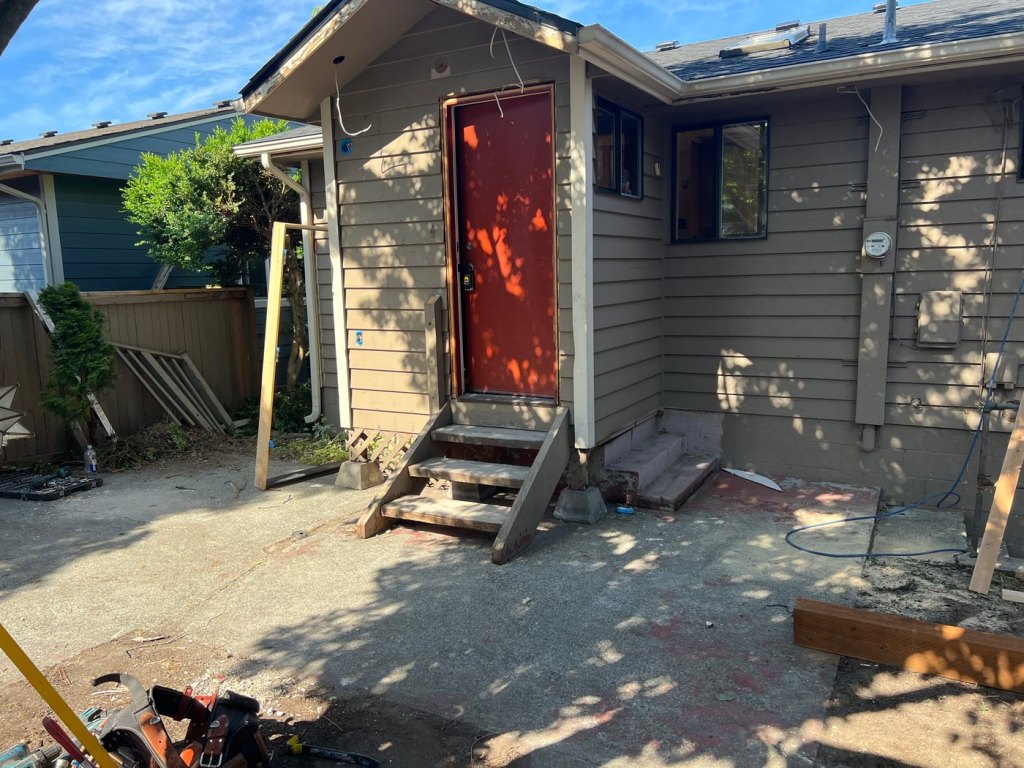 The door hung and closed at the same entry, with the old wood steps still in place and lumber stacked by the fence.